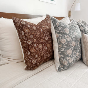 A cozy bed with white and beige bedding is adorned with decorative throw pillows. The pillows feature intricate patterns, including one in burgundy with floral designs and another in a muted green floral print, adding a touch of elegance to the setup.