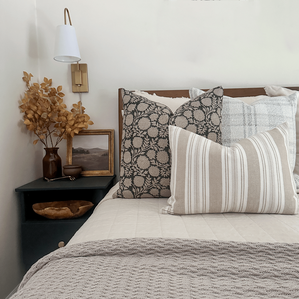 Callum pillow cover on the left and Tobias pillow on the right with the Odin lumbar pillow cover in front. Set on a neatly made bed with floral decor on side table and wall-mounted gold lamp.