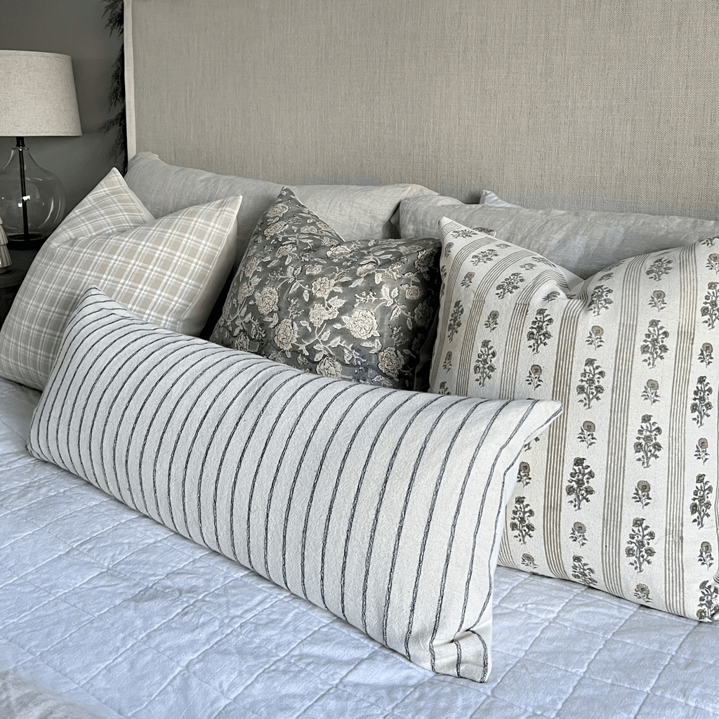 A neatly arranged bed features a variety of decorative pillows, including the Lenora Pillow Cover in natural linen. Several throw pillows boast different patterns, such as block-printed florals, stripes, and checks in soft neutral tones, all set against a light gray headboard.