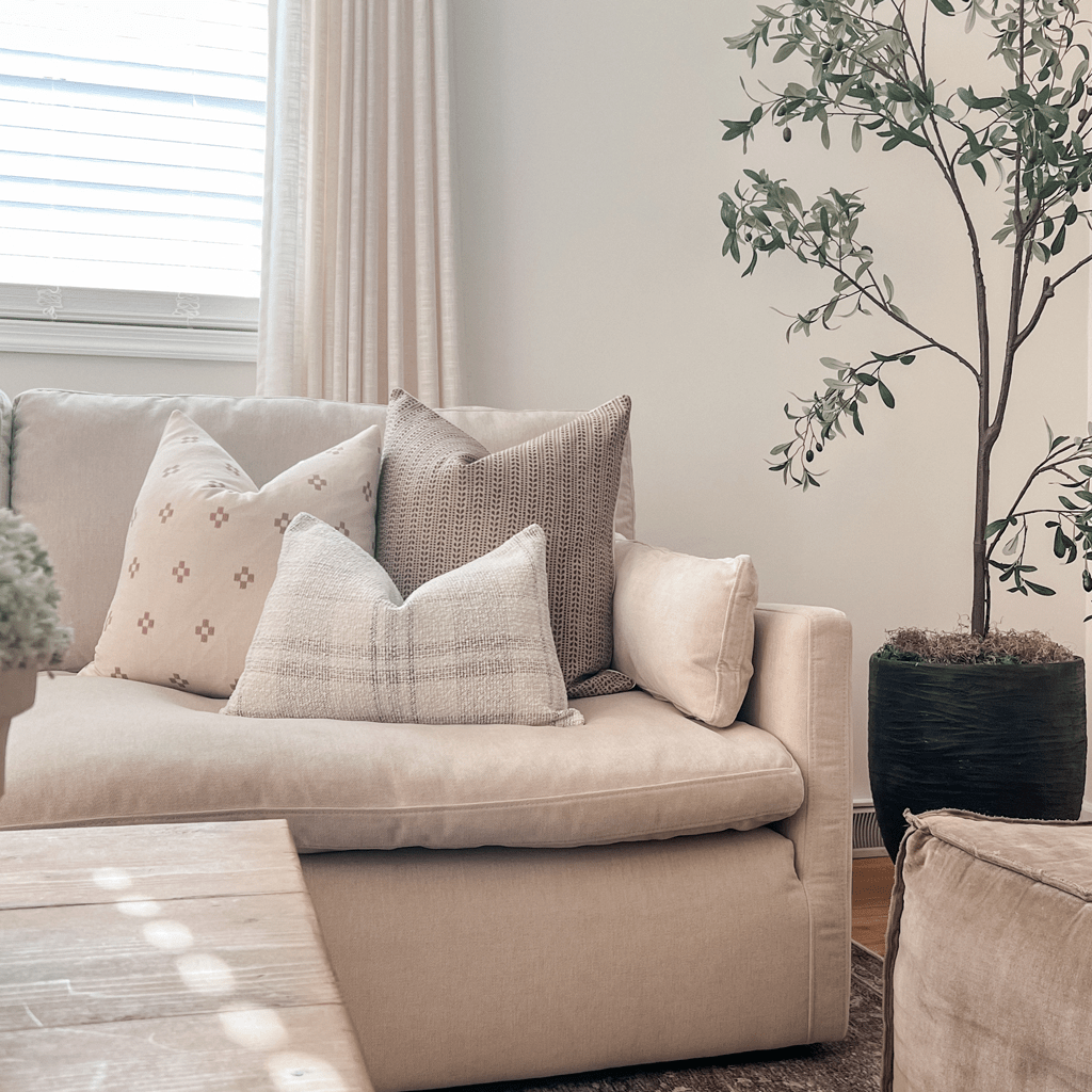 A cozy living room scene features a light beige sofa adorned with a Tobias Lumbar Pillow Cover and two other decorative pillows in various patterns. A tall potted plant stands nearby, and sunlight filters through white blinds and light curtains. A wooden table is in the foreground.