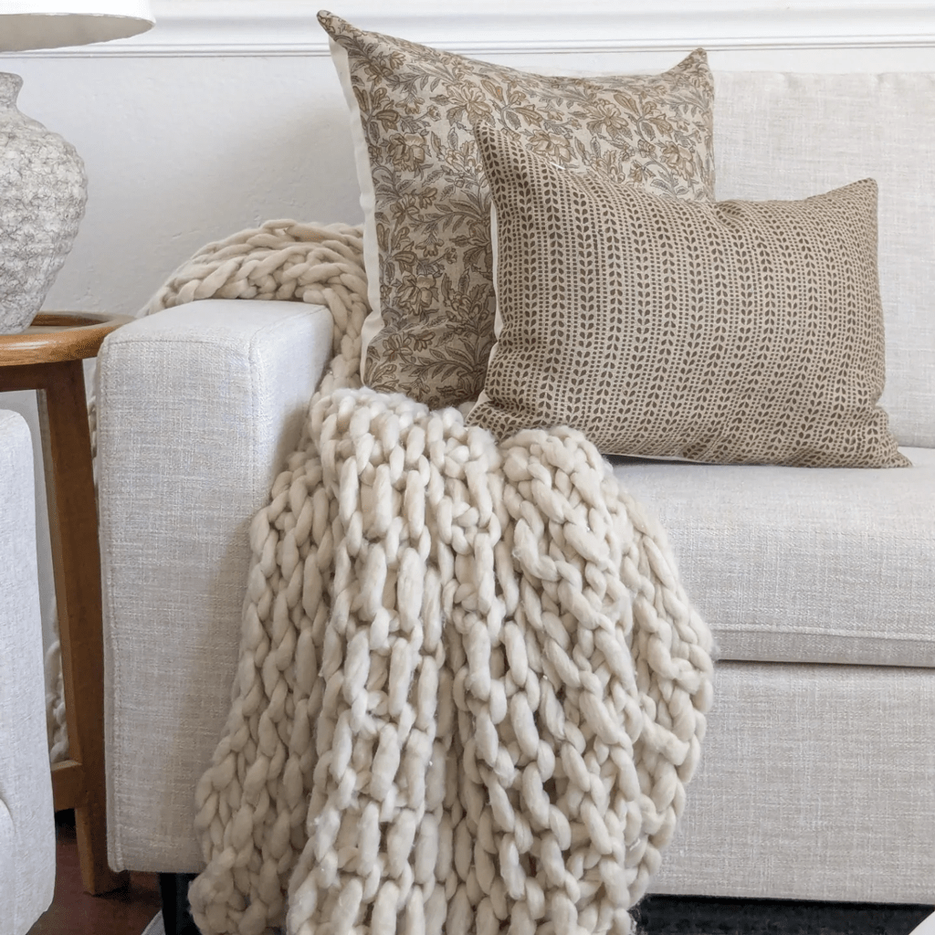 A cozy scene with a beige sofa adorned with two patterned pillow covers and a chunky knit cream blanket draped over the arm. A wooden side table with a textured vase is partially visible to the left, adding to the handwoven charm of this inviting space.