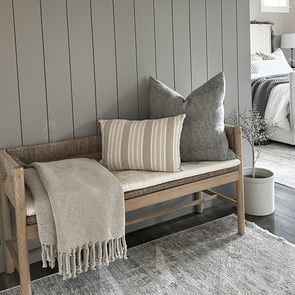 A cozy wooden bench with a woven backrest is adorned with striped pillows alongside a floral print pillow cover. A textured throw blanket is draped over it. Next to the bench, a potted plant stands on the floor, set against the backdrop of a neutral-toned bedroom.