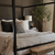 A cozy bedroom with a black four-poster bed adorned with checkered and patterned pillows, alongside a blanket featuring wooly texture. A wooden bench sits at the foot of the bed. A charcoal grey windowpane frames the scene, while a potted plant and table lamp sit on the bedside table.