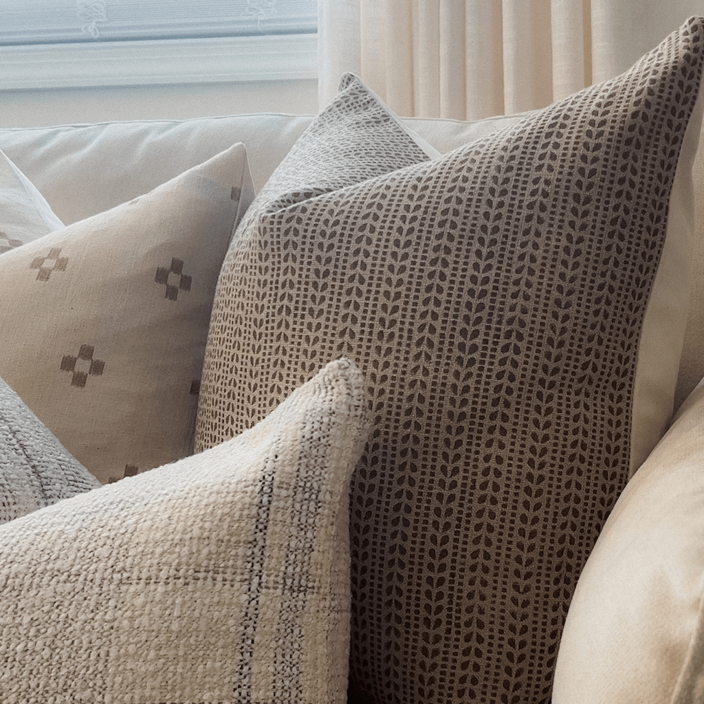 A cozy arrangement of four decorative pillows adorns the couch, including a brown one with a textured design and an Ester Pillow Cover featuring a rust floral motif. Nearby, beige curtains made from natural flax linen frame the scene, adding warmth and elegance to the setting.