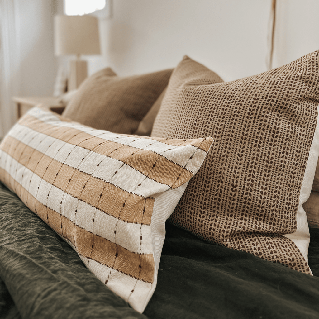 A set of pillows on a bed, featuring Colin and Finn's mix of striped and patterned designs in beige and brown tones. The bedding is dark green, with an Ester Pillow Cover adding a rust floral motif, while a lamp glows softly in the background.