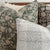 Close-up of a group of decorative throw pillows on a bed. One pillow has a green floral pattern, another features a beige and dark brown botanical design, and a third showcases a textured, light gray and white plaid pattern. The background includes a fluffy white pillow.