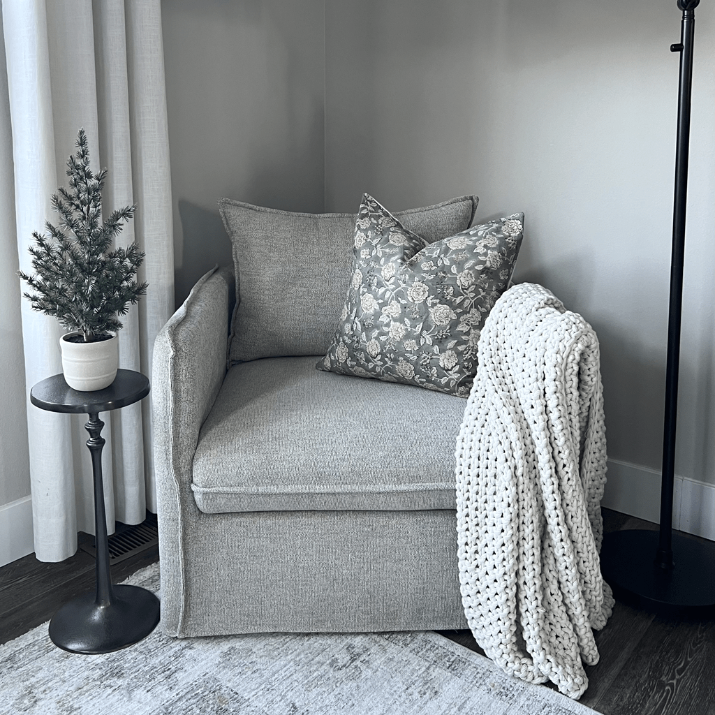 A cozy corner with a gray armchair featuring floral pillows and a chunky knit blanket. A potted evergreen rests on a round black side table. The Lenora Pillow Covers add charm, while light curtains adorn the window and a floor lamp stands nearby, casting a soft glow.