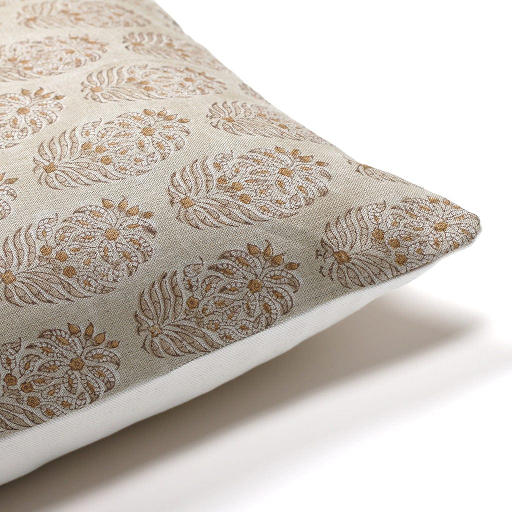 A close-up of a beige pillow with an intricate, repeated paisley pattern in gold and white. The pillow is resting on a white surface, highlighting the delicate design and texture of the fabric.