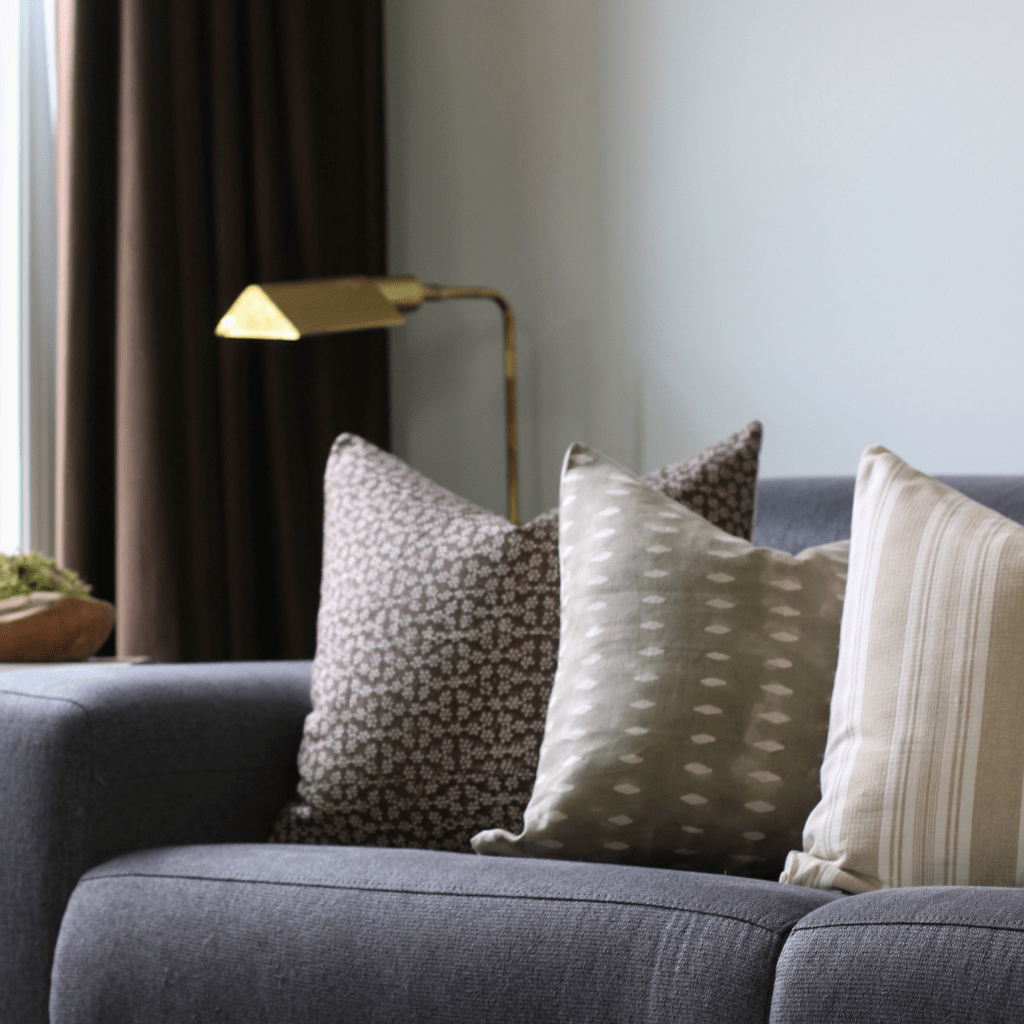 Neville Mocha, Lee, and Odin throw pillows from Colin + Finn on a blue sofa with a gold lamp and brown curtains in the background. 