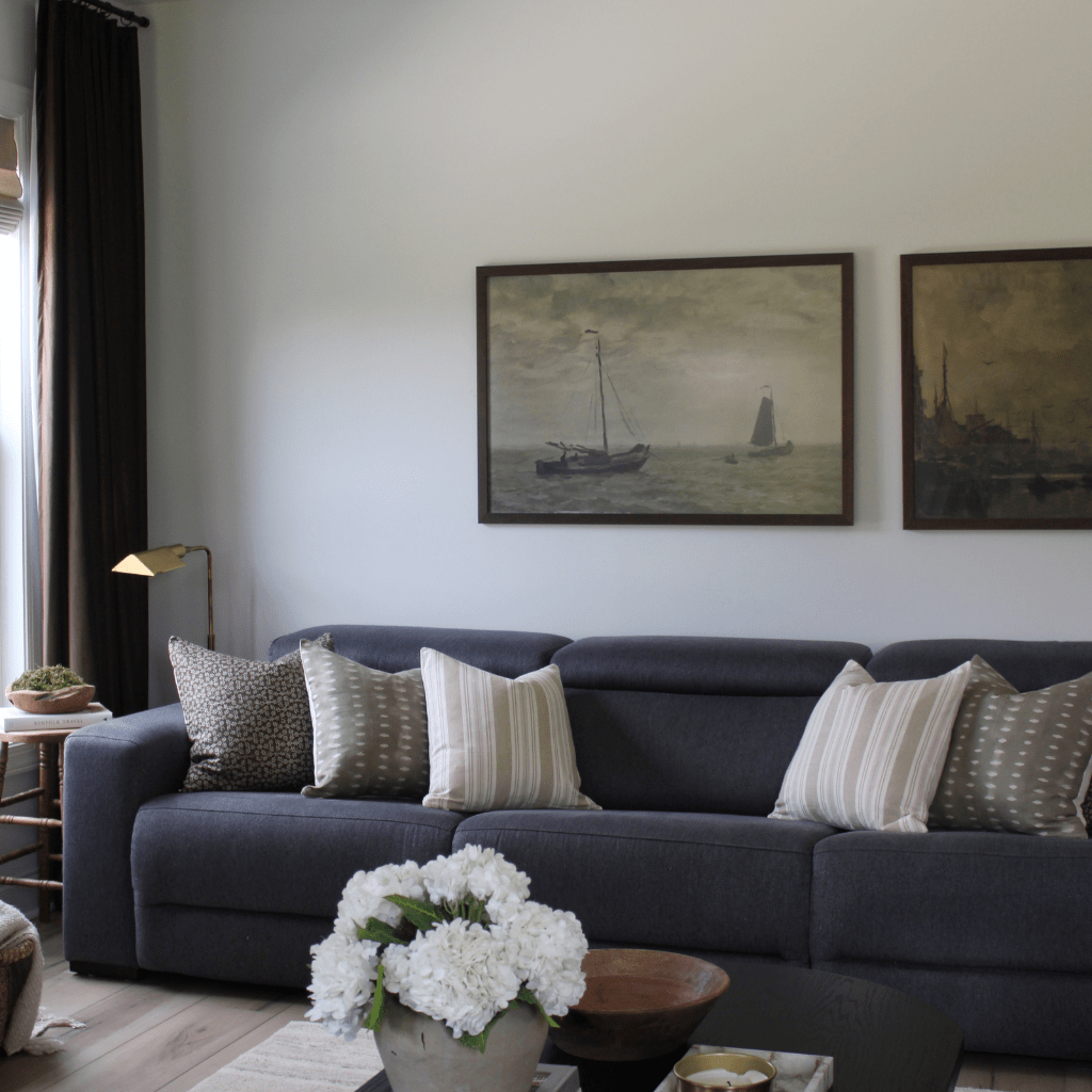 A living room with Neville Mocha, Lee, and Odin throw pillows from Colin + Finn on a blue sofa with a gold lamp and brown curtains in the background. Two nautical paintings are on the wall and white flowers and a bowl are in the foreground of the photo.